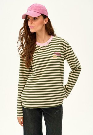 Kobieta ubrana w zielono-biały, w paski, długi rękaw z napisem „Sweet Escape”, różowy kołnierzyk, różowa czapka i czarne spodnie, patrząca na bok.