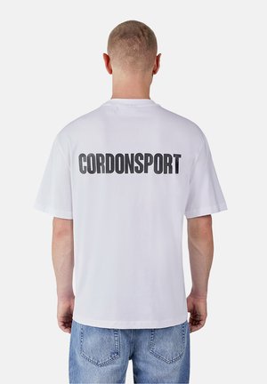 Mann trägt ein weißes T-Shirt mit "CORDONSPORT" in fetten schwarzen Buchstaben auf dem Rücken, kombiniert mit blauen Denim-Jeans.