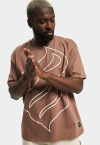 T-shirt in cotone marrone a maniche corte, con un prominente design astratto bianco. Include un piccolo etichetta nera all'orlo.