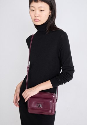 Kobieta w czarnym golfie trzymająca małą bordową torebkę na ramię z srebrnym logo CK na przedniej kieszeni.