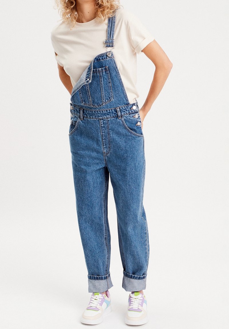 Pantalones de mezclilla en azul medio con tirantes ajustables, botones laterales y puños doblados, combinados con zapatillas blancas con acentos en tonos pastel.