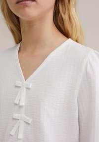 Vit texturerad blus med V-ringning och tre tygband vid fronten. Har samlade ärmar och mjukt, lätt material.