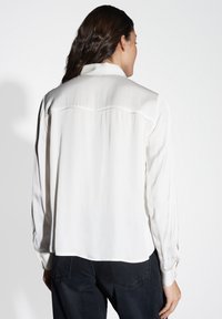 Blusa bianca a maniche lunghe realizzata in tessuto liscio, con colletto classico, polsini abbottonati e un dettaglio di spalline sulla schiena.