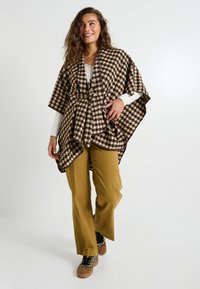 Poncho à carreaux marron et crème avec un lien à l'avant, porté sur une chemise blanche à manches longues et un pantalon vert olive, associé à des chaussures à motifs.