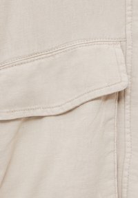Beige fabric close-up featuring a pocket with a flap, fine stitching, and a smooth texture. Subtle variations in color and texture visible.