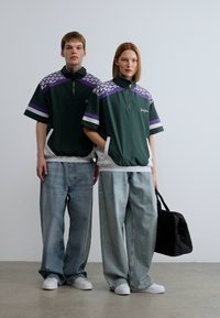 Πράσινο φούτερ με φερμουάρ στο μισό μήκος και μοτίβα σε μωβ και λευκό γεωμετρικό σχέδιο, σε συνδυασμό με φαρδιά τζιν σε ανοιχτή ντενίλ απόχρωση και λευκά αθλητικά παπούτσια. Συμπεριλαμβάνεται μαύρη τσάντα tote.