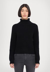 Schwarzer Strickpullover mit Rollkragen, gerippter Textur, langen Ärmeln und einer verkürzten Silhouette. Kombiniert mit dunkelgrauen Jeans.