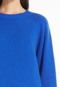 Kobieta ubrana w jasnoniebieski sweter z prążkowanego materiału, z długimi rękawami i okrągłym dekoltem, na białym tle.