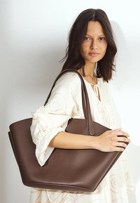 Sac en cuir marron avec une texture lisse et un grand haut ouvert, doté de longues manches robustes et d'un design minimaliste.