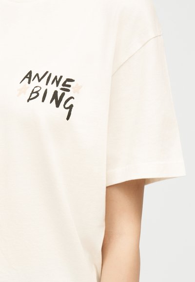 Off-white short-sleeve shirt with black handwritten text "ANINE BING" and two small beige stars on the chest, worn by a person.