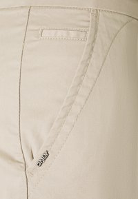 Béžové kalhoty z hladké látky, s boční kapsou s prošitým detailem a malou kovovou štítkem s logem na pase.