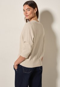 Beige gestrickter Pullover mit geripptem Muster, Dreiviertelärmeln und rundem Ausschnitt, kombiniert mit dunkelblauen Jeanshosen.