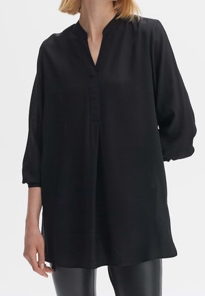 Frau trägt eine lockere schwarze Tunika mit langem Arm, V-Ausschnitt und plissierten Details, kombiniert mit schwarzen Lederhosen vor weißem Hintergrund.