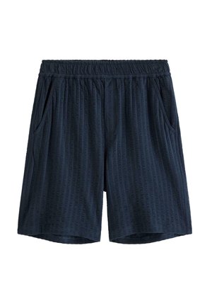 Dreng i marineblå skjorte med knapper og korte ærmer samt matchende shorts, iført en brun kasket bagvendt og sorte sandaler, stående på en ensfarvet baggrund.