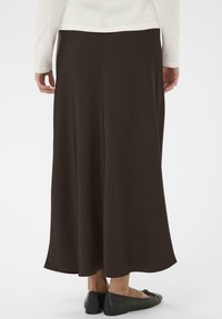 Bruine, vloeiende maxi rok gemaakt van een soepele stof, met een volle silhoët en een zoom die net boven de enkels valt. Zwarte ballerina's zijn zichtbaar.
