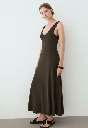 Kobieta w bezrękawowej, długiej sukience w ciemnym oliwkowym kolorze z głębokim dekoltem w kształcie litery V, w połączeniu z czarnymi sandałami i brązowymi bransoletkami, stoi na tle gładkiej ściany.