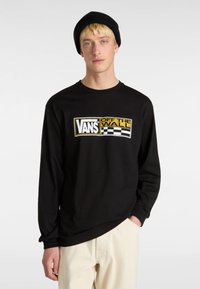 Schwarzes Langarmshirt mit einem grafischen Logo in Gelb und Weiß. Das Material wirkt weich und hat eine lässige Passform, kombiniert mit hellen Hosen.