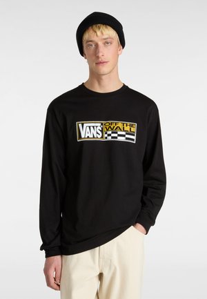 Black long-sleeve shirt featuring a graphic logo in yellow and white. Material appears soft with a relaxed fit, paired with light-colored pants.