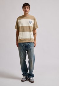 Brun og creme stribet t-shirt med korte ærmer og broderet logo detaljer, parret med lyseblå denim jeans og grå sneakers.