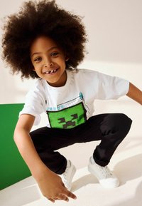 Maglietta grafica bianca con un design di faccia pixelata verde, pantaloni neri e sneakers bianche, indossata da un bambino con capelli ricci.