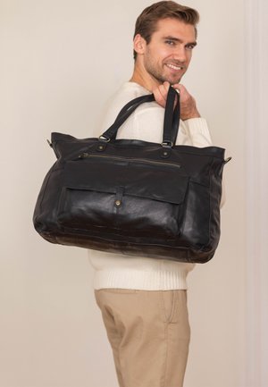 Black leather bag with dual handles, zippered top, and front pocket. Smooth texture with subtle stitching accents. Large, rectangular shape.