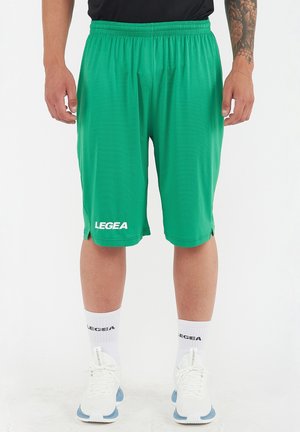 Uomo che indossa pantaloncini sportivi verdi, calzini bianchi e scarpe da ginnastica bianche, in piedi davanti a uno sfondo bianco uniforme.