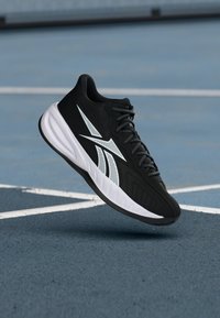 Zapatilla deportiva en blanco y negro con suela gruesa, flotando sobre una cancha exterior azul con líneas blancas.