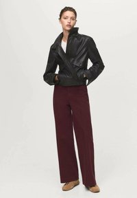 Giubbotto di pelle nera con colletto alto, dettagli con bottoni e design asimmetrico abbinato a pantaloni larghi bordeaux.