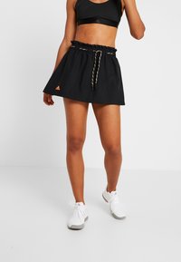 Kvinna iklädd svart träningskjol med dragsko och crop top, vita sneakers, stående mot vit bakgrund.