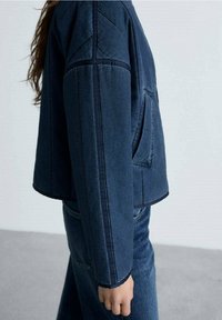 Veste en denim foncé avec une coupe carrée, coutures apparentes et poche latérale. Associée à un pantalon en denim foncé assorti pour un look coordonné.