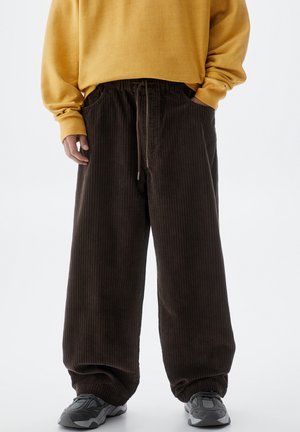 Person iført løse, mørkebrune kordfløjlsbukser med snøre, sennepsgul sweatshirt og grå sneakers på en hvid baggrund.