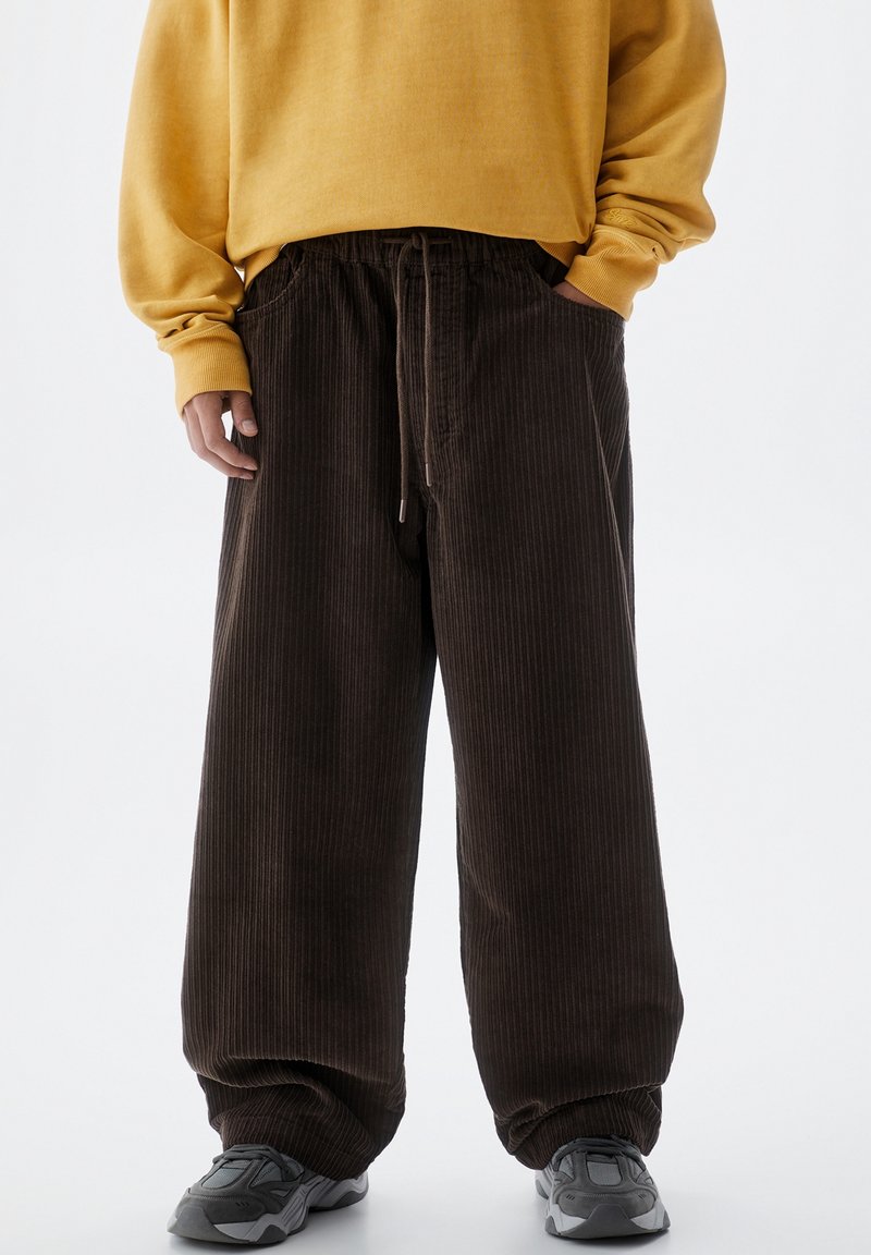 Personne portant un pantalon ample en velours côtelé marron foncé avec cordon, un sweat-shirt jaune moutarde et des baskets grises sur fond blanc.