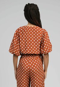 Combinaison marron à pois avec des manches bouffantes, une coupe décontractée, une taille élastique et une texture de tissu lisse. Pois blancs visibles.