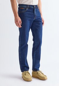 Homme portant un jean droit bleu foncé et des chaussures en daim marron clair à lacets, debout sur un fond blanc.