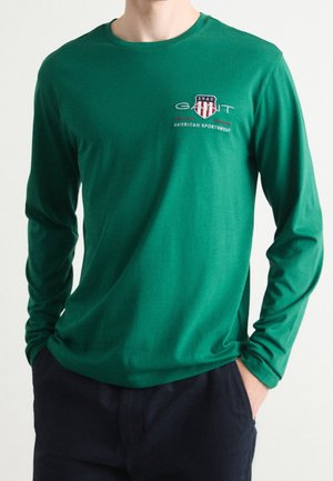 Mężczyzna w zielonej koszuli z długim rękawem z logo "GANT 1949 American Sportswear" na piersi, z rękami w kieszeniach, w ciemnych spodniach.