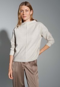 Helle beige Strickjacke mit hohem Kragen, überschnittenen Schultern und einer Mittelnaht-Detail, kombiniert mit glatten, braunen Satin-Hosen.
