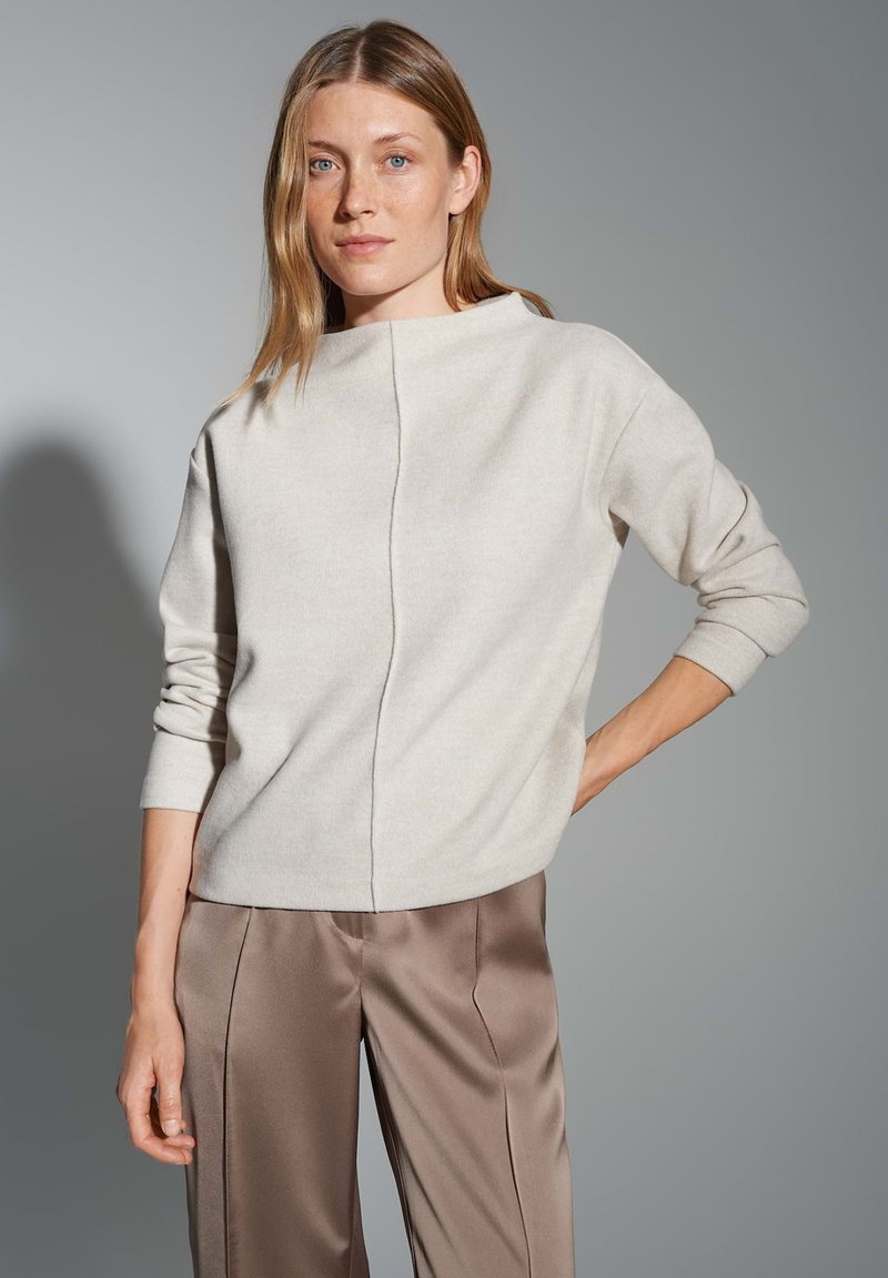 Helle beige Strickjacke mit hohem Kragen, überschnittenen Schultern und einer Mittelnaht-Detail, kombiniert mit glatten, braunen Satin-Hosen.