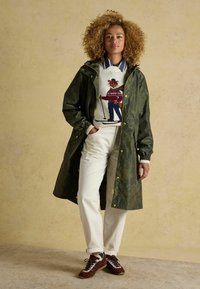 Moteris su garbanotais plaukais, vilkinti žalią gėlių rašto lietpaltį, megztinį su slidinėjimo lapės piešiniu, baltas kelnes ir rudas sportines batus, stovi prieš beige spalvos sieną.