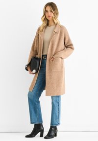 Beige wool blend long coat with a relaxed fit, front pockets, and notched collar, paired with blue straight-leg jeans and black heeled ankle boots.