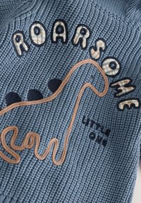 Tissu bleu tricoté présentant le texte "ROARSOME" et un motif abstrait de dinosaure en marron et marine, avec trois points marines au-dessus.