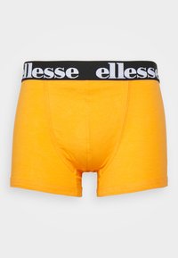Dzelteni kokvilnas bokseršorti ar melnu elastīgu jostu, uz kuras ir balts "ellesse" zīmols, un plakano šuvju apdare, kas nodrošina gludu apdari.