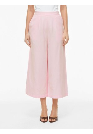 Žena má na sebe ružové krátke nohavice s vysokým pásom a širokými nohavicami, bočnými vreckami a hnedými otvorenými opätkami na bielom pozadí.