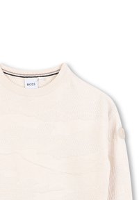 Maglione lavorato a maglia beige con un design testurizzato che presenta ondulazioni sottili. Collo a giro e dettaglio del pulsante con logo sulla manica sinistra.