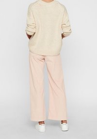 En person ses bakifrån, iförd en ljusbeige tröja, ljusrosa vid byxor och vita sneakers. Bakgrunden är helt grå.