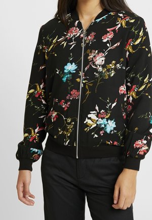 Femme portant une veste noire à fermeture zippée avec un motif floral multicolore, des poignets et un ourlet côtelés, associée à un pantalon noir.