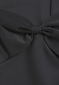 Svart tyg med en slät textur har en stor rosettdetalj i mitten, säkrad med ett matchande band. Den övergripande designen är draperad.