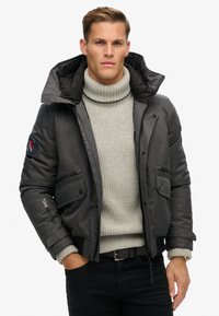 Grijze gewatteerde jas met een afneembare capuchon, voorzakken en logo-detail op de mouw. Draagt over een crèmekleurige turtleneck trui.