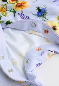 Gros plan sur un tissu avec un imprimé floral coloré et des boutons-pression en métal sur un fond lavande clair.