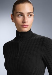 Svart ribbat stickat linne med hög krage och halvdämpad dragkedja, med en figursydd design som framhäver textur och subtila linjer.
