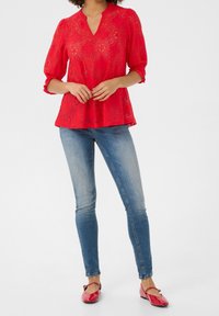 Top in pizzo rosso con scollatura a V e maniche a sbuffo, abbinato a jeans skinny blu e ballerine rosse con un dettaglio decorativo.
